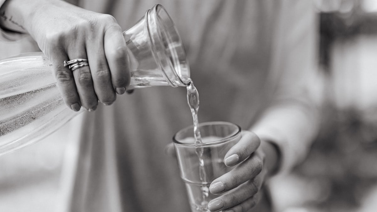 A close-up view of a woman pouring water from a carafe into a glass, highlighting hands and jewelry.